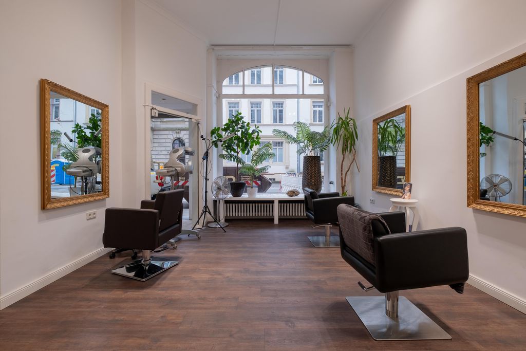 Innenraum eines Friseursalons mit zwei Friseurstühlen, großen Spiegeln an den Wänden und Grünpflanzen vor einem großen Fenster.