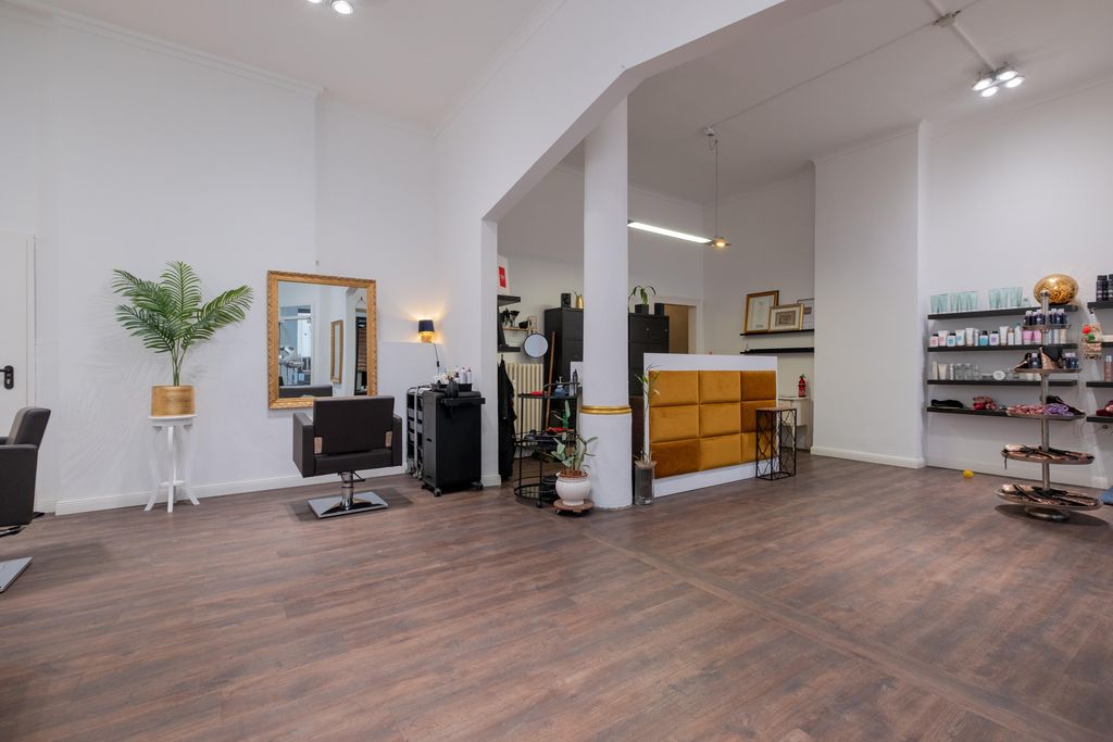 Ein leerer Friseursalon mit zwei Friseurstühlen, einem großen Spiegel, einer Pflanze in einem goldenen Topf und einem Empfangstresen mit braunen Polstern.