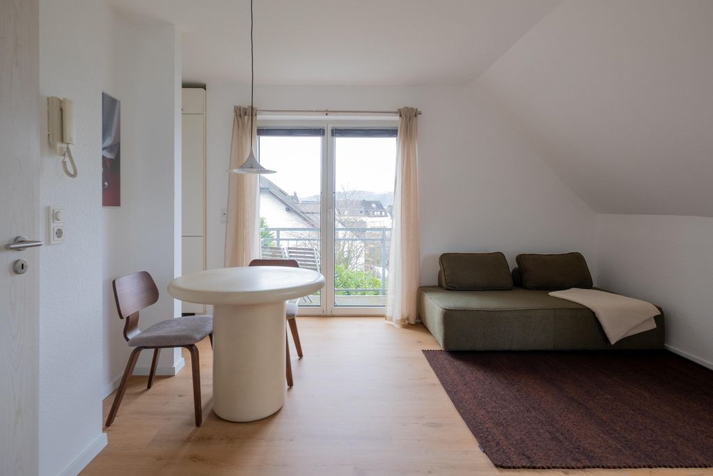 Heller Raum mit rundem weißen Esstisch, zwei Stühlen und einem grünen Sofa mit Kissen und Decke neben einem großen Fenster mit Blick auf Häuser und Bäume.