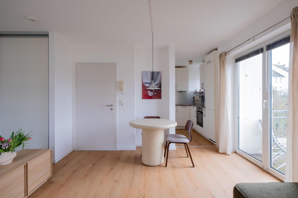 Ein moderner, heller Essbereich mit einem runden weißen Tisch und zwei braunen Stühlen vor einer weißen Tür, mit einer Küche im Hintergrund und großen Fenstern zu einem Balkon.