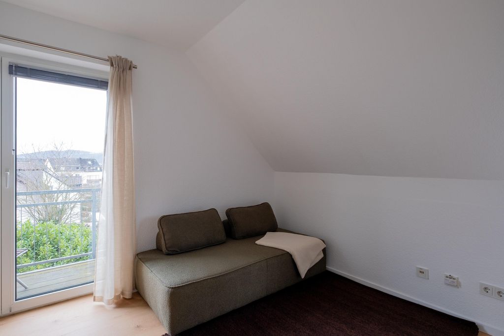 Ein schlichter Raum mit einer grauen Couch, zwei Rückenpolstern und einem hellen Überwurf neben einer Balkontür mit halbtransparenten Vorhängen.