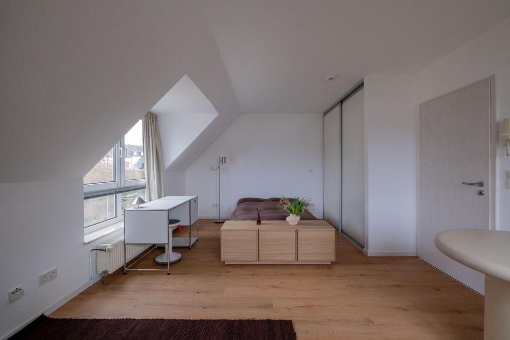Schlafzimmer unter einem Schrägdach mit großem Fenster, Schreibtisch, Doppelbett mit brauner Bettdecke und Holzkommode mit Pflanzenvasen.