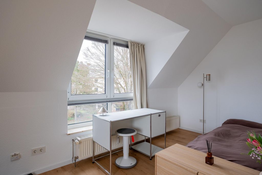 Helles Schlafzimmer mit einer Schräge, großem Fenster, weißem Schreibtisch mit Hocker, Stehlampe und einem Bett mit braunem Überwurf.