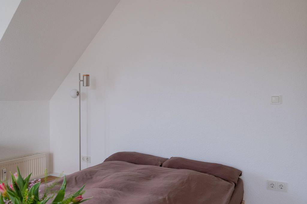 Ein schlichtes Schlafzimmer mit einem großen Bett, braunen Kissen und einer weißen Wand im Hintergrund. Im Vordergrund sind Pflanzblätter sichtbar.
