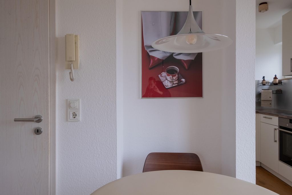 Ein runder Esstisch mit einem Holzstuhl vor einer weißen Wand mit einem modernen Hängelicht und einem Bild mit einer Tasse Kaffee und einem Tablett darauf.