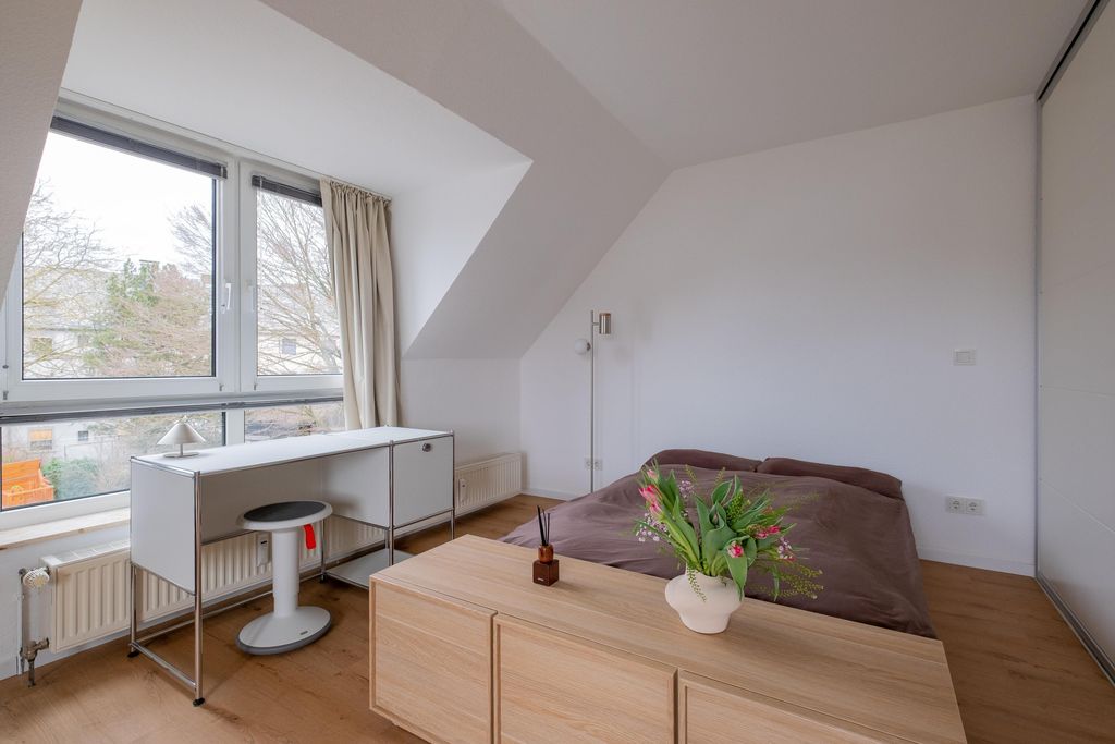 Ein helles Schlafzimmer mit einem großen Fenster, einem einfachen Schreibtisch mit Hocker, einem Doppelbett mit brauner Bettdecke und einer Holzkommode mit Vase und Blumen darauf.