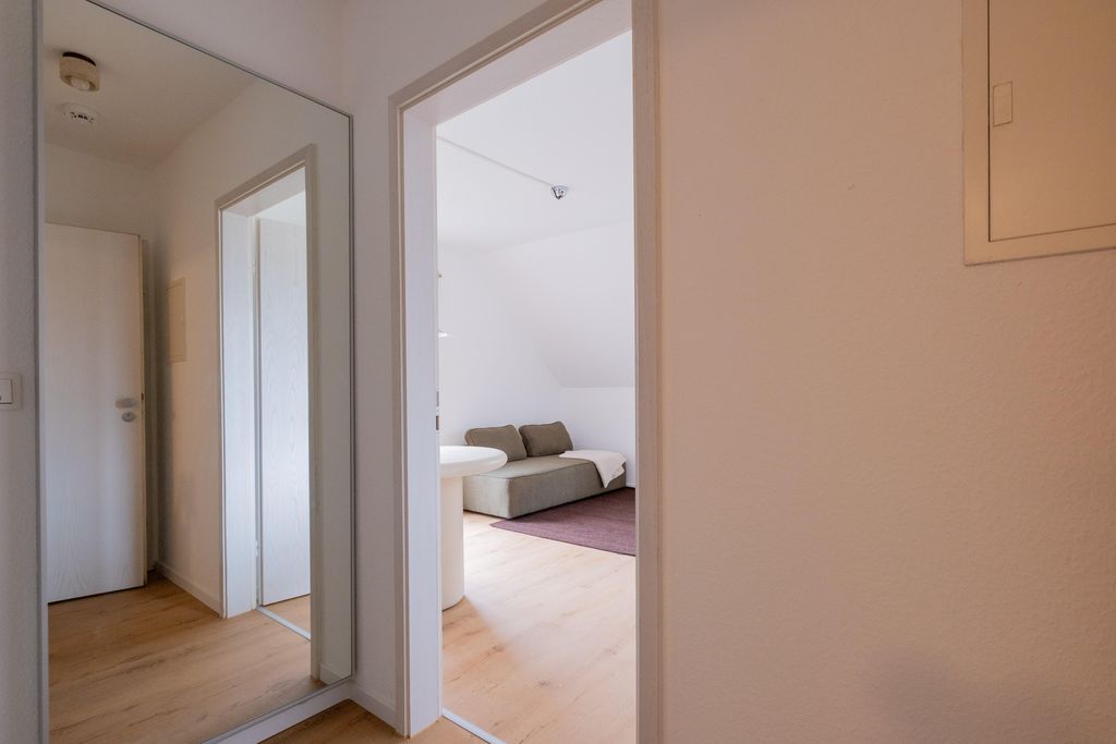 Blick durch eine offene Zimmertür in ein Wohnzimmer mit hellen Holzböden, einem grauen Sofa und einem runden weißen Tisch.