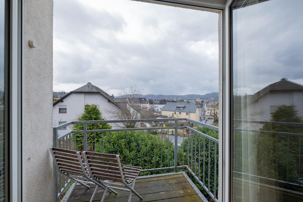 Balkon mit zwei Holzstühlen und Geländer, Blick auf eine bewachsene Wohngegend unter bewölktem Himmel.