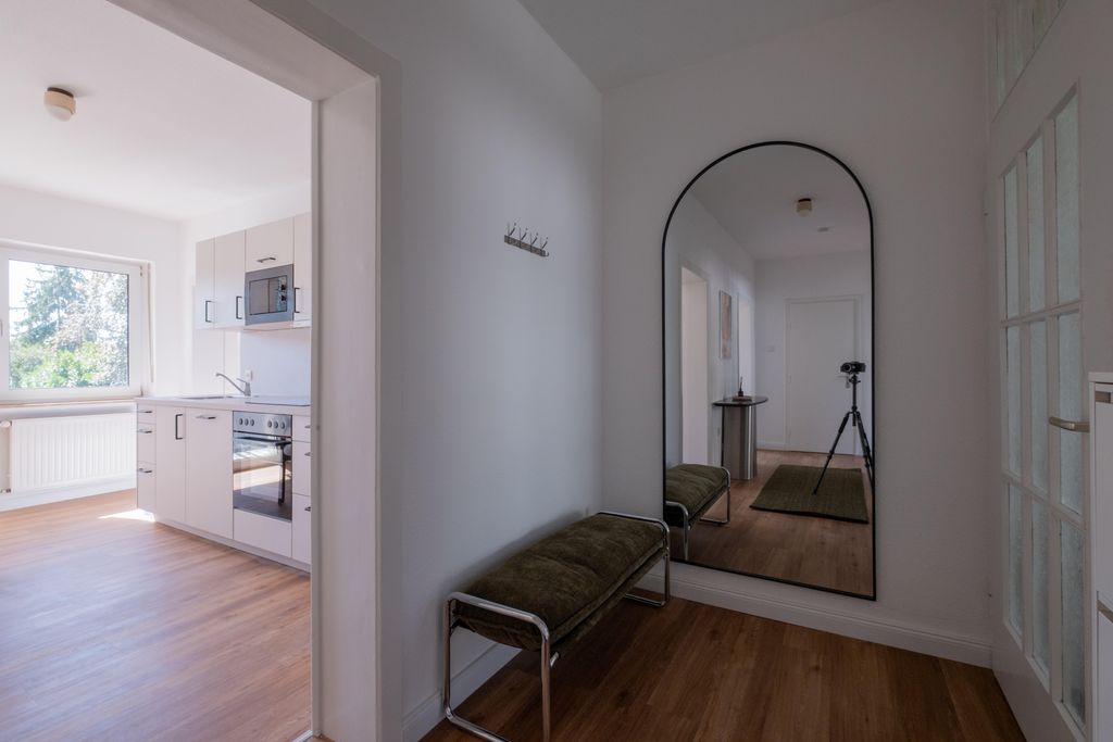 Eingangsbereich mit einem schmalen Sitzbank mit grünem Polster, einem großen Wandspiegel mit schwarzem Rahmen und einem Blick in eine helle Küche mit weißen Schränken und Holzfußboden.