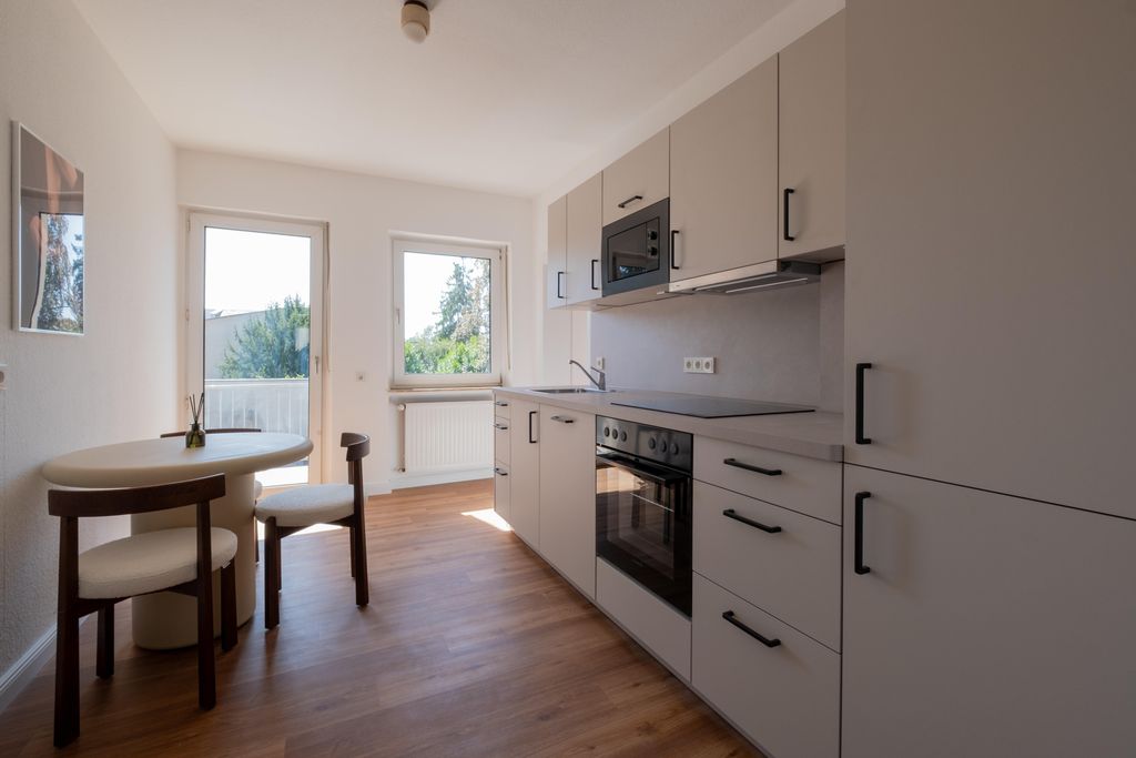 Helle moderne Einbauküche mit weiß-mattem Mobiliar, einem Kochfeld, Backofen und Mikrowelle sowie einem kleinen runden Tisch mit zwei Stühlen vor einem Fenster und einer Tür.