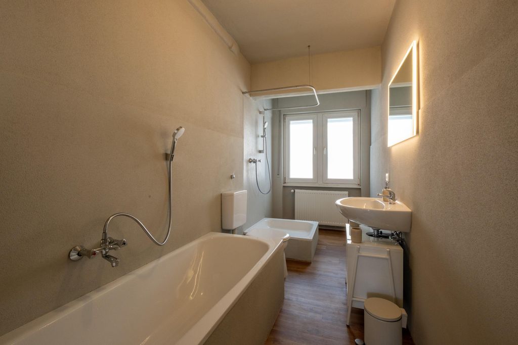 Schlicht eingerichtetes Badezimmer mit Badewanne, Waschbecken, beleuchtetem Spiegel und Fenster an der Rückseite.