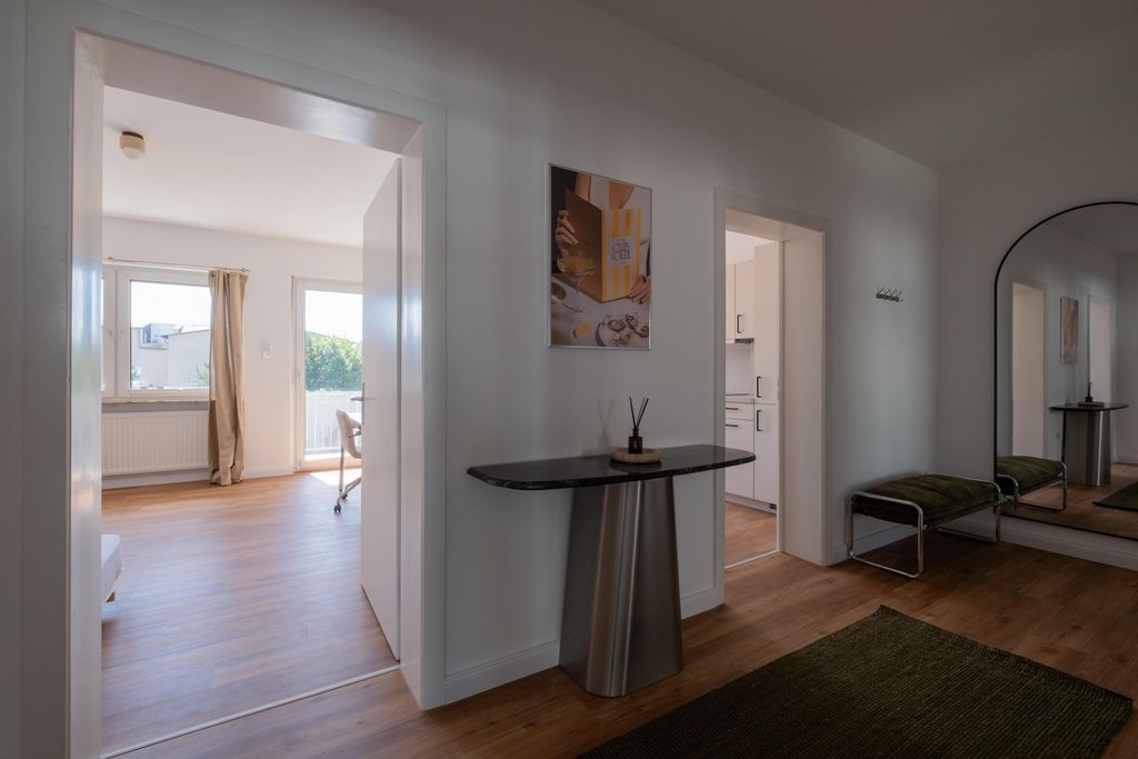 Heller Hausflur mit braunem Holzboden, kleiner schwarzer Tisch mit Dekoration, grünem Teppich, Bank rechts und großem Spiegel an der Wand.