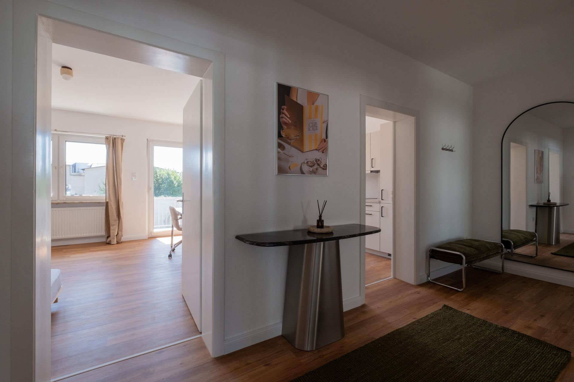 Heller Hausflur mit braunem Holzboden, kleiner schwarzer Tisch mit Dekoration, grünem Teppich, Bank rechts und großem Spiegel an der Wand.