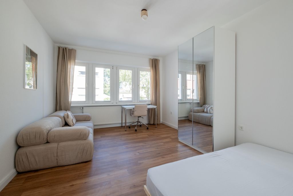 Helles Zimmer mit einem Doppelbett, beige Couch mit Kissen, einem Schreibtisch mit Stuhl und einem großen Kleiderschrank mit Spiegeltüren vor einem Fenster mit Vorhängen.