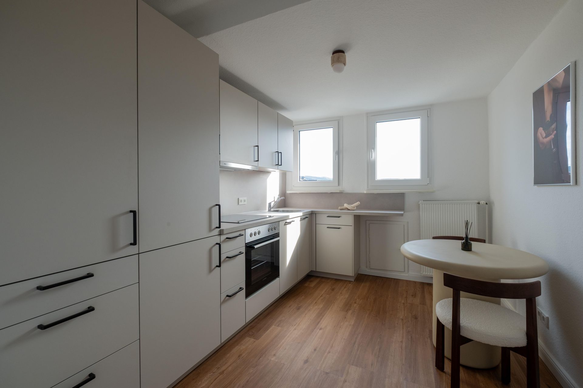 Moderne, minimalistisch eingerichtete Küche mit weißen Schränken, Holzboden, zwei Fenstern und einem kleinen runden Esstisch mit zwei Stühlen.