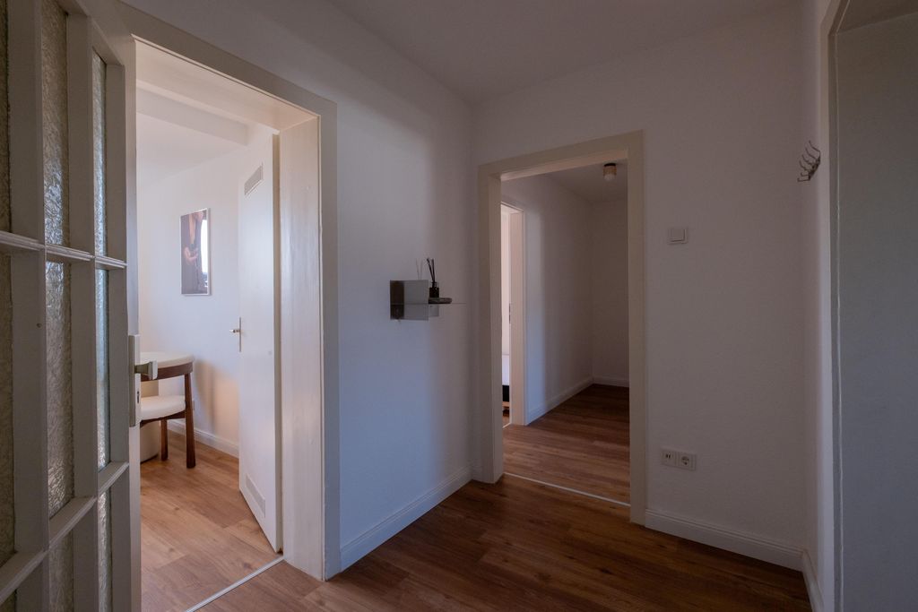 Blick in einen Innenflur mit weißen Wänden und Holzboden, zwei offene Türen führen in angrenzende Zimmer mit weißen Möbeln.