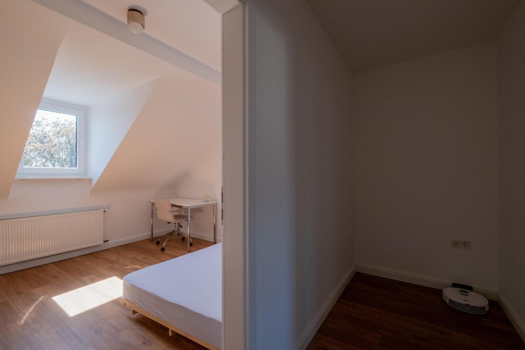 Zimmer mit Schräge, das in zwei Bereiche unterteilt ist: links ein Bereich mit einem Bett, Stuhl und Schreibtisch vor einem Fenster; rechts ein dunklerer Bereich mit einem Saugroboter auf dem Boden.
