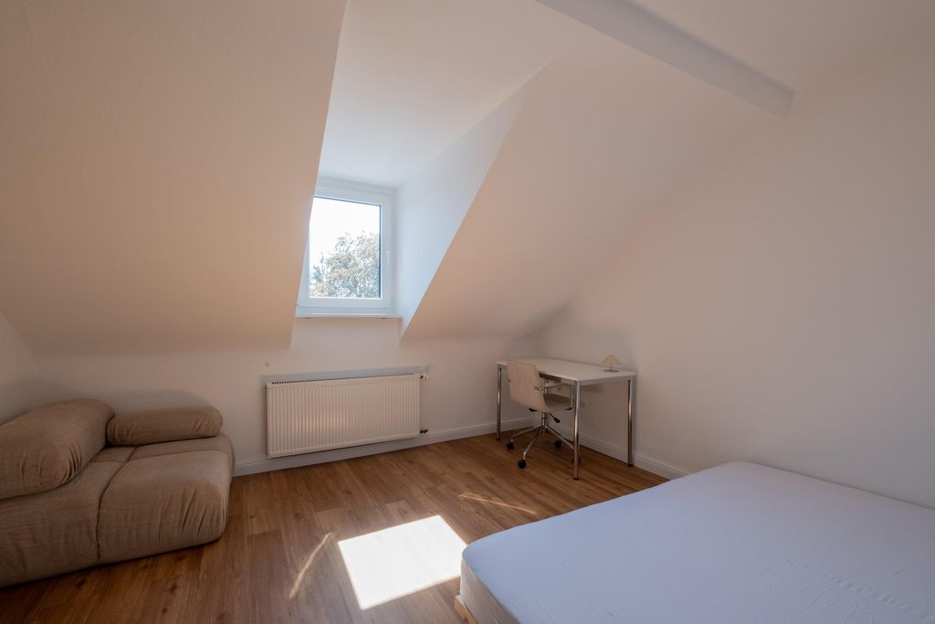 Helles, minimalistisches Schlafzimmer mit schräger Decke, einem kleinen Fenster, weißem Schreibtisch mit Stuhl, einem beigen Sessel und einem Bett mit weißer Matratze auf Holzboden.