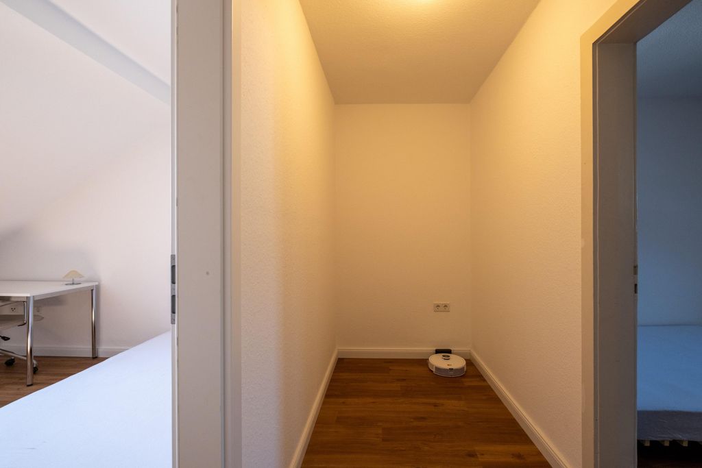 Ein kleiner Raum mit weißen Wänden und einem Holzboden, in dem ein weißer Saugroboter an der Wand steht. Links und rechts sind offene Türen zu angrenzenden Zimmern sichtbar.