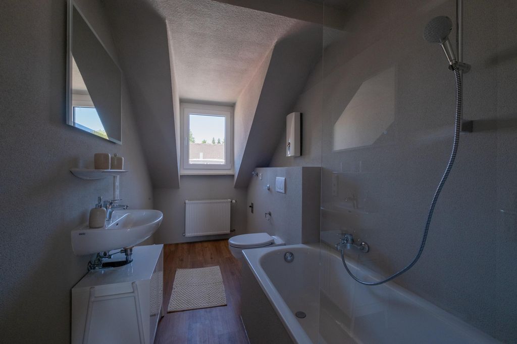Ein Badezimmer mit einer Badewanne, einem bidetähnlichen Toilettenaufsatz, einem Waschbecken auf einem weißen Unterschrank, einem Fenster unter einer Schräge und einem Holzfußboden mit einem kleinen Teppich.
