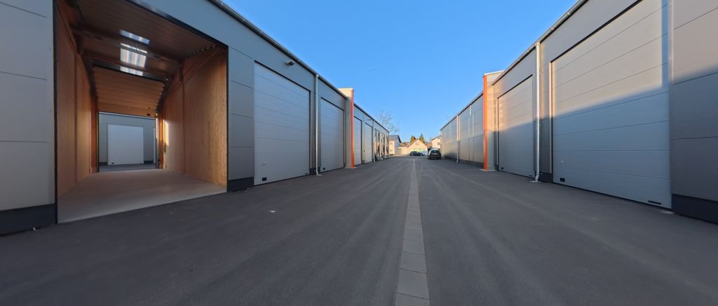 Eine breite, asphaltierte Straße zwischen zwei Reihen moderner Industriehallen mit großen, geschlossenen Toren unter klarem blauem Himmel.
