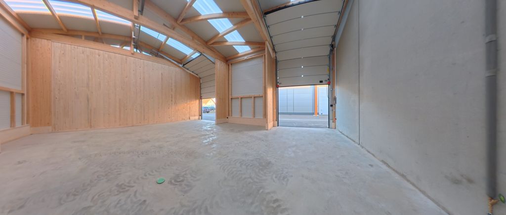 Innenansicht einer leerstehenden, neu gebauten Halle mit Holzwänden und großem geöffnetem Tor, das auf einen Parkplatz hinausführt.