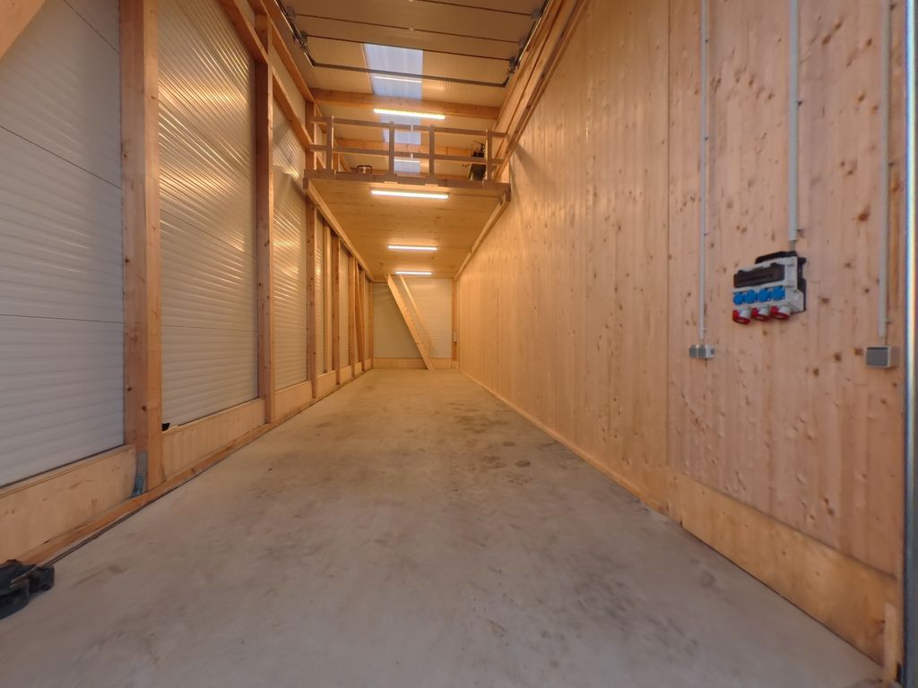 Ein langer, schmaler Lagerraum mit Holzverkleidungen an den Wänden und drei geschlossenen Rolltoren auf der linken Seite, beleuchtet von Deckenlampen, mit einem Treppenaufgang am Ende des Raums.