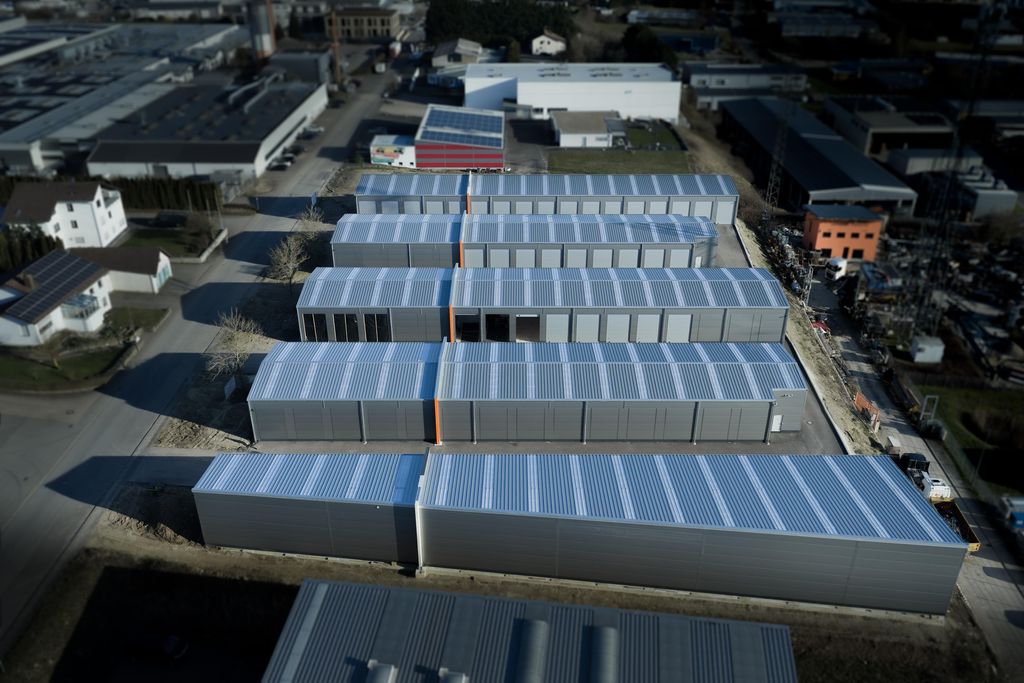 Luftaufnahme eines Industriegebiets mit mehreren langen, grauen Lagerhallen mit blaugrauen Dächern, die in Reihen angeordnet sind.