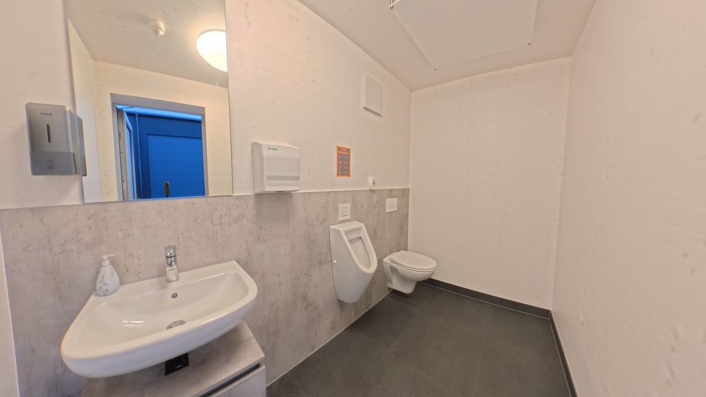 Ein sauberer öffentlicher Waschraum mit einem Waschbecken, Urinal und einer Toilette, weiße Wände und grauer Bodenbelag.