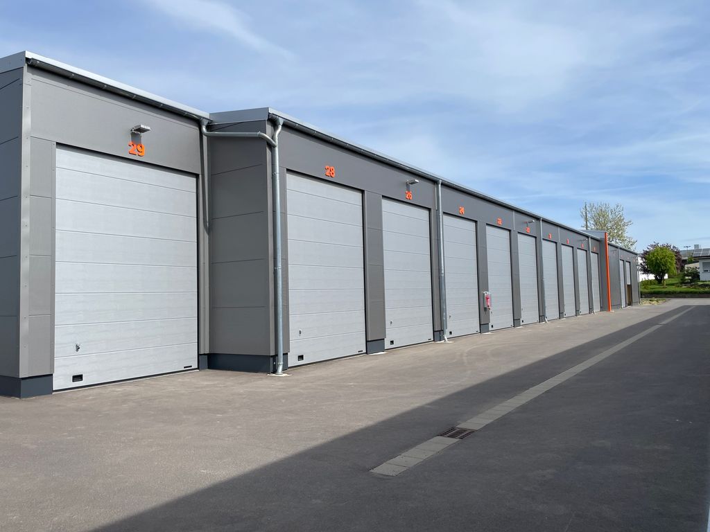 Reihe von geschlossenen, grauen Industriegaragentoren mit orangen Zahlen 29 bis 21, aufgenommen bei sonnigem Himmel.