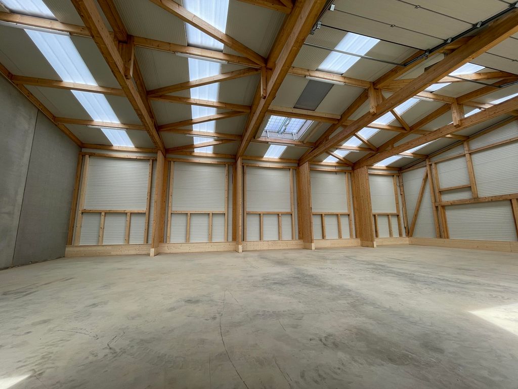 Leerer Lagerraum mit Betonboden, Holzträgern und mehreren geschlossenen Rolltoren entlang der Wände, beleuchtet von Tageslicht durch Dachfenster.