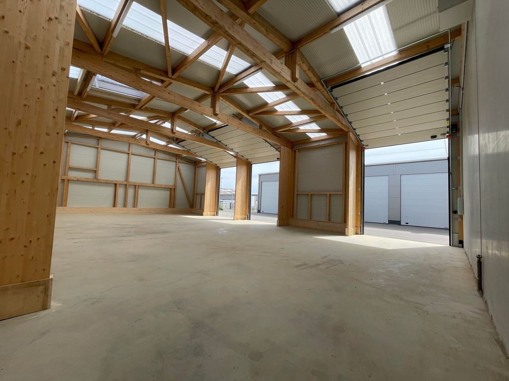 Leere Halle mit Holzrahmenkonstruktion und offenem Garagentor, die natürliches Licht durch die transluzenten Dächer hereinlässt.