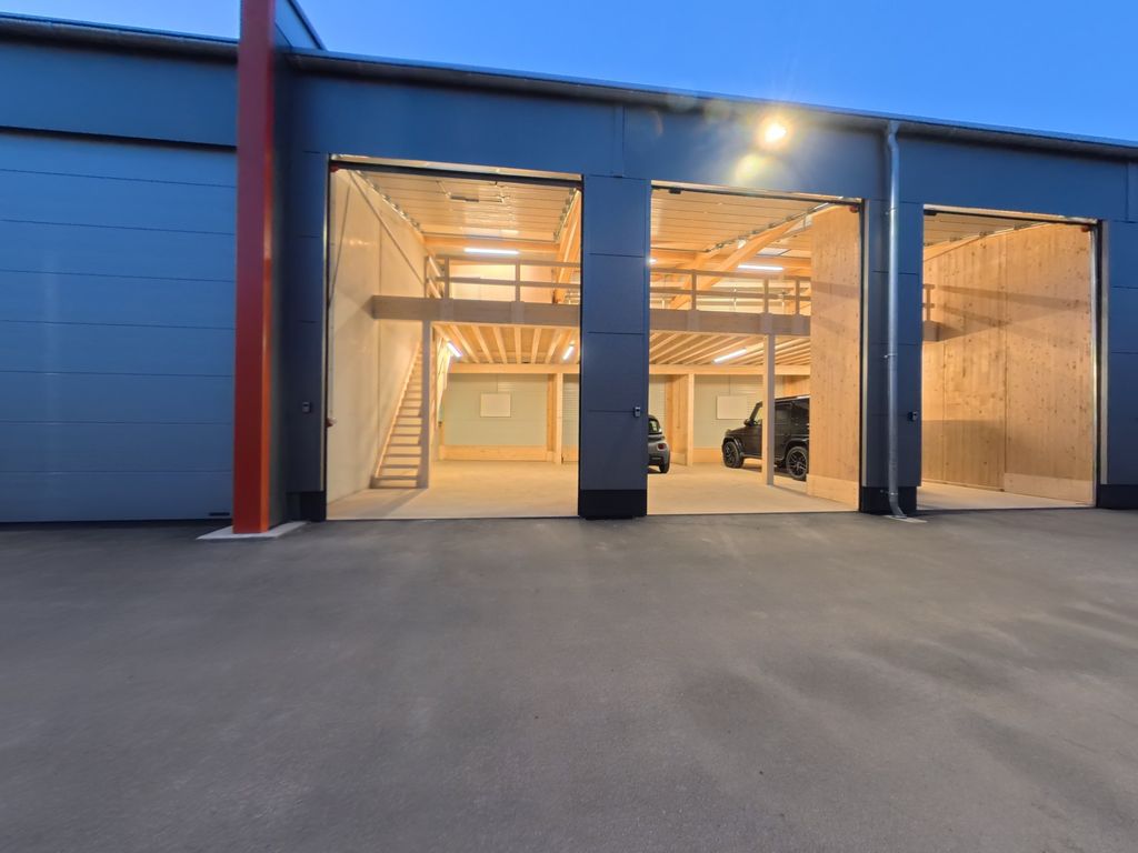 Eine moderne Lagerhalle oder Garage mit großen offenen Toren, beleuchtet bei Dämmerung, mit sichtbaren Fahrzeugen und einer Holzstruktur im Inneren.