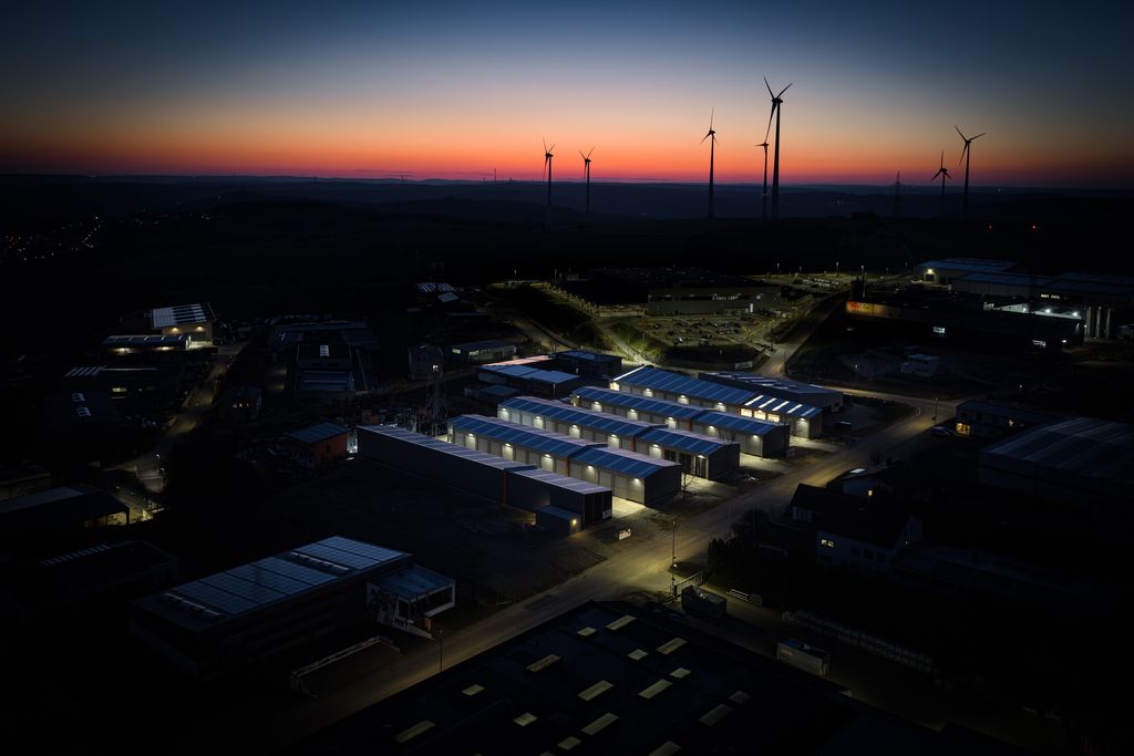 Industriegelände mit beleuchteten Lagerhallen bei Sonnenuntergang, im Hintergrund sind mehrere Windräder an einem Hügel zu sehen.