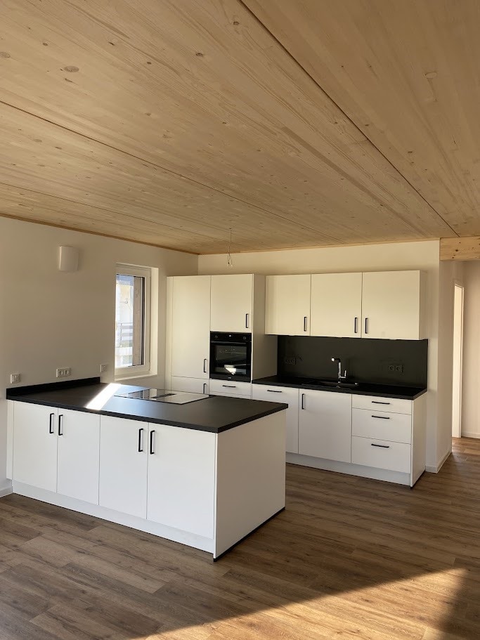 Moderne Küche mit weiß lackierten Schränken, schwarzen Arbeitsflächen und Holzdecke in einem hellen Raum mit Fenster.
