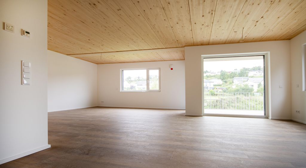 Großer leerer Raum mit hellen Wänden, Holzdecke und breiter Fensterfront mit Blick auf eine grüne Landschaft.