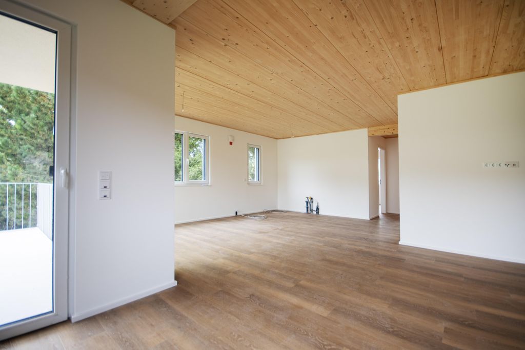 Leerer Raum mit hellen weißen Wänden, großen Fenstern und einer Tür zu einem Balkon, Holzboden und Holzdecke.