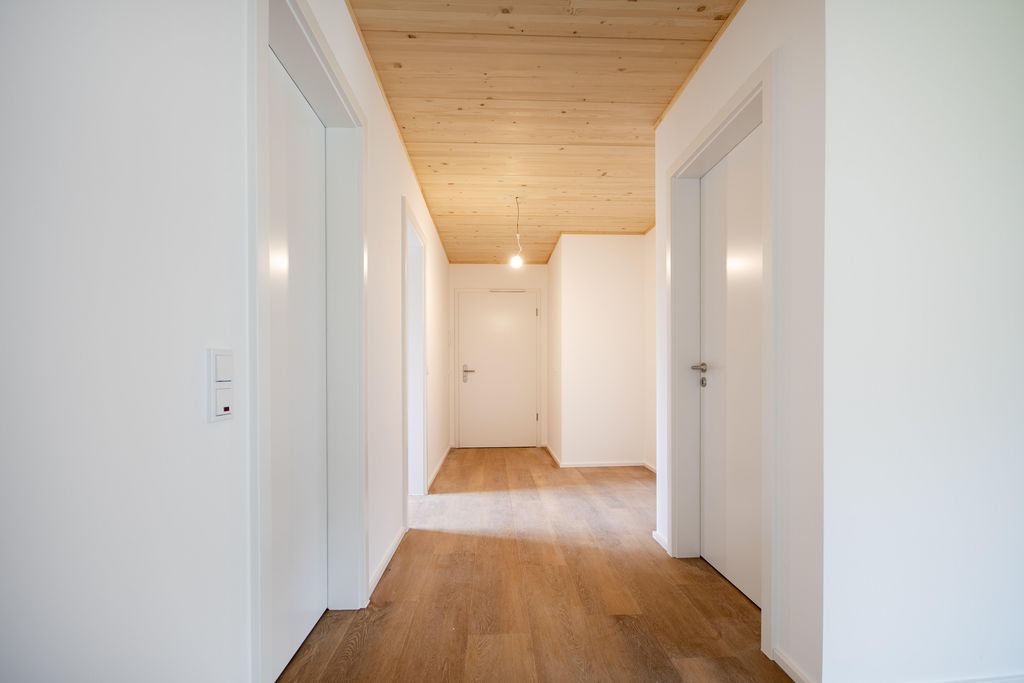 Hell beleuchteter Flur mit weißen Wänden, Holzboden und Holzdecke, drei sichtbare weiße Türen und eine Pendelleuchte an der Decke.