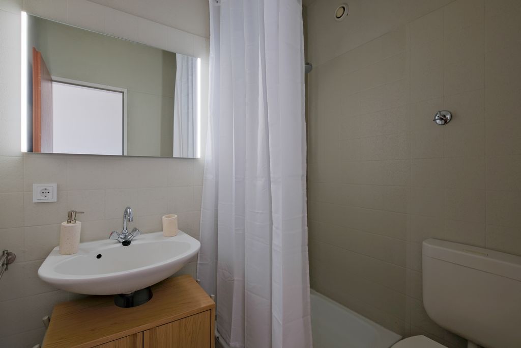 Kleines Badezimmer mit weißem Waschbecken, Holzunterschrank, beleuchtetem Spiegel, weißem Duschvorhang und WC mit grauen Fliesen.