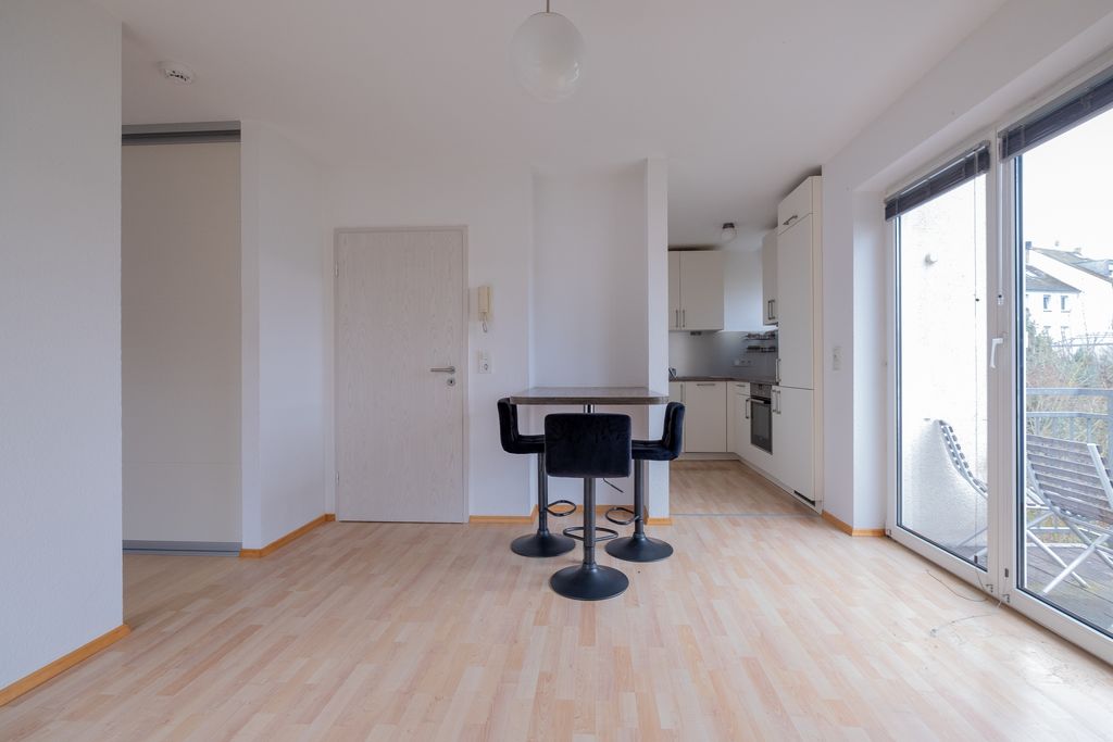 Moderner, leerer Wohnraum mit hell holzfarbenem Laminatboden, einer kleinen Bar mit drei schwarzen Barhockern in der Mitte und einer offenen Küche im Hintergrund. Große Fenster mit Blick auf einen Balkon und eine hell beleuchtete Wand mit einer geschlossenen Tür.