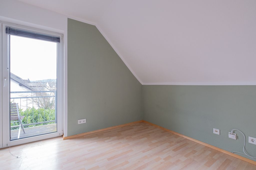 Leerer Raum mit Holzparkettboden, grüner Wand und großer Glastür mit Blick auf eine Terrasse und Garten.