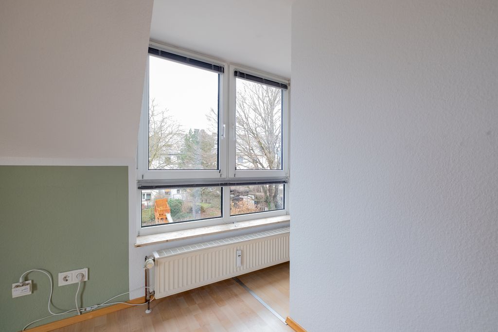 Zimmer mit schräger Decke, einem großen Fenster mit weißen Rahmen, darunter ein Heizkörper, und einem grünen Wandabschnitt neben weißen Wänden.