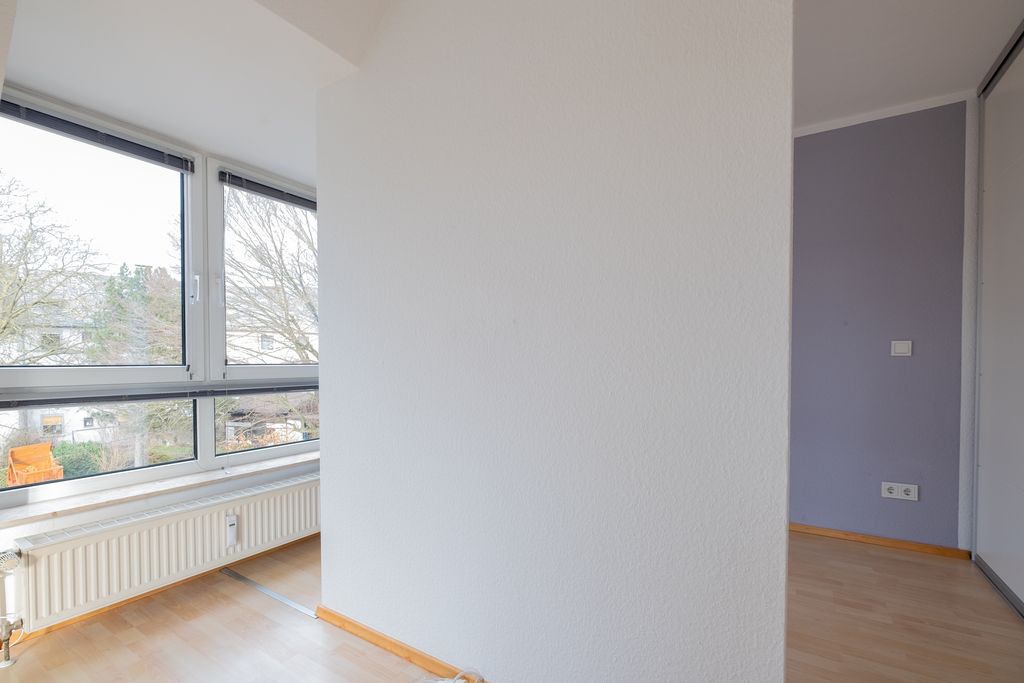 Leerer Raum mit hellen Holzböden und großen Fenstern, die Tageslicht hereinlassen, mit Sicht auf Bäume und Wohnhäuser außen.