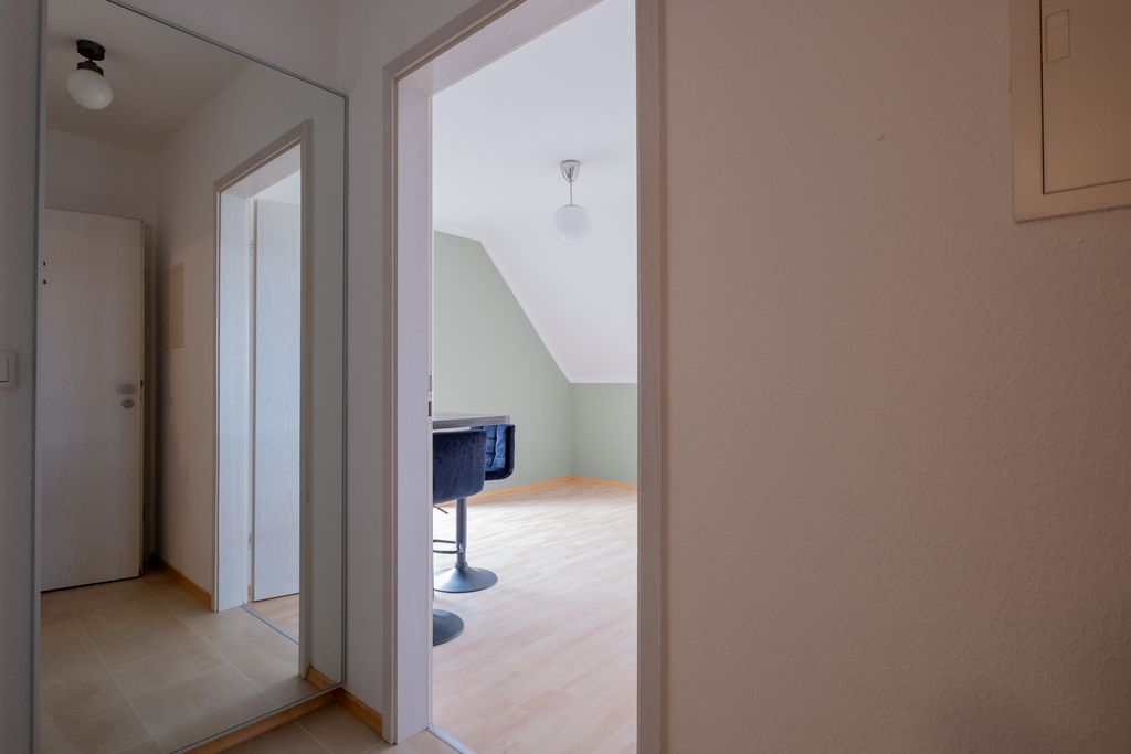 Flur eines Hauses mit Blick in ein Zimmer mit Parkettboden, eingeschränktem Dachschrägenbereich und einem schwarzen Stuhl an einem Fensterplatz.