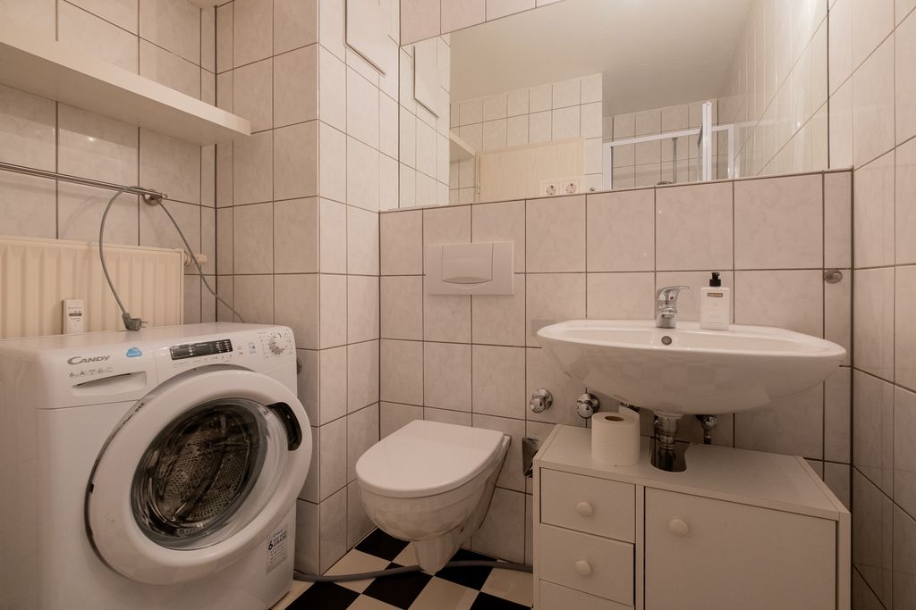 Ein kleines Badezimmer mit einer Waschmaschine, einer Wandtoilette und einem Waschbecken auf einem Unterschrank, weiß gekachelte Wände und schwarz-weiß karierten Boden.