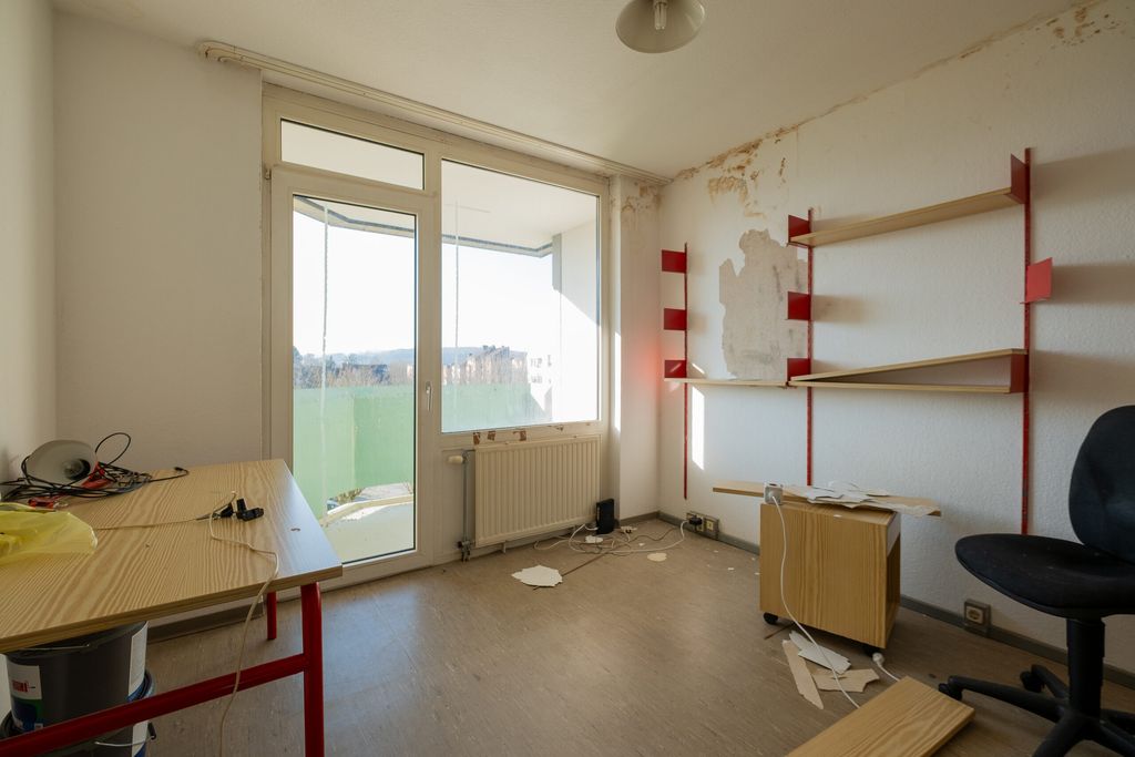Leerer Raum mit Holzregalen mit roten Halterungen an der weißen Wand, beschädigter Wandfarbe, einem Tisch mit Kabeln und einem Stuhl. Große Glastür führt zu einem Balkon mit Blick ins Freie.
