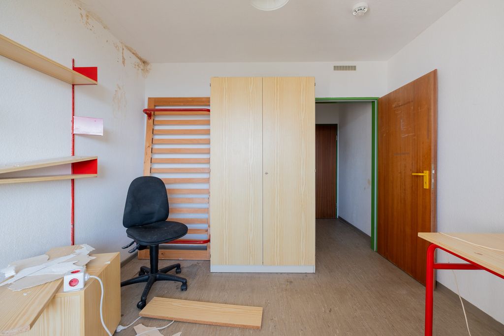 Sehr einfacher Raum mit einem großen Holzkleiderschrank, einem schwarzen Bürostuhl, einem offenen braunen Türrahmen, Regalen an der linken Wand und einem Tisch mit roten Beinen rechts.