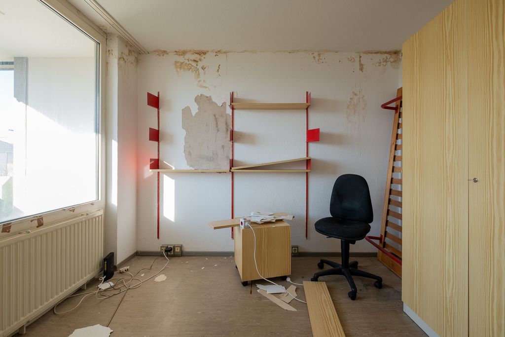 Blick in ein leeres und vernachlässigtes Zimmer mit einem beschädigten weißen Wandbereich, Regal mit roten Halterungen, Bürostuhl, kleinem Holzschrank und Holzmöbeln, die in gutem Zustand sind.