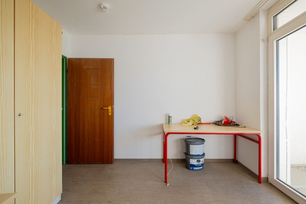 Leerer Raum mit einem Holztisch mit roten Beinen, auf dem verschiedene Werkzeuge und ein gelbes Tuch liegen, daneben mehrere Farbeimer, eine geöffnete Holztür und ein großer Fenstertürbereich, der viel Licht hereinlässt.