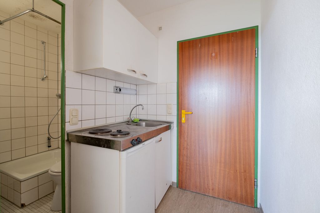 Kleine Küchenzeile mit Herdplatte, Spüle und weißen Schränken neben einer Holztür mit gelbem Griff in einem hellen Raum.